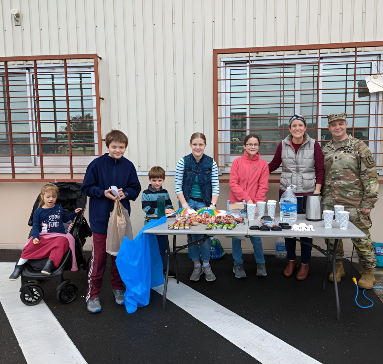 A man in a U.S. Army uniform, his wife and their 5 children of various ages stand behind a table full of snacks.