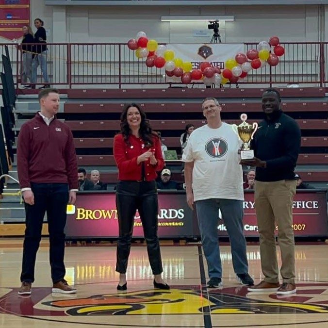 Four people on a basketball court with one person holding and displaying a trophy.
