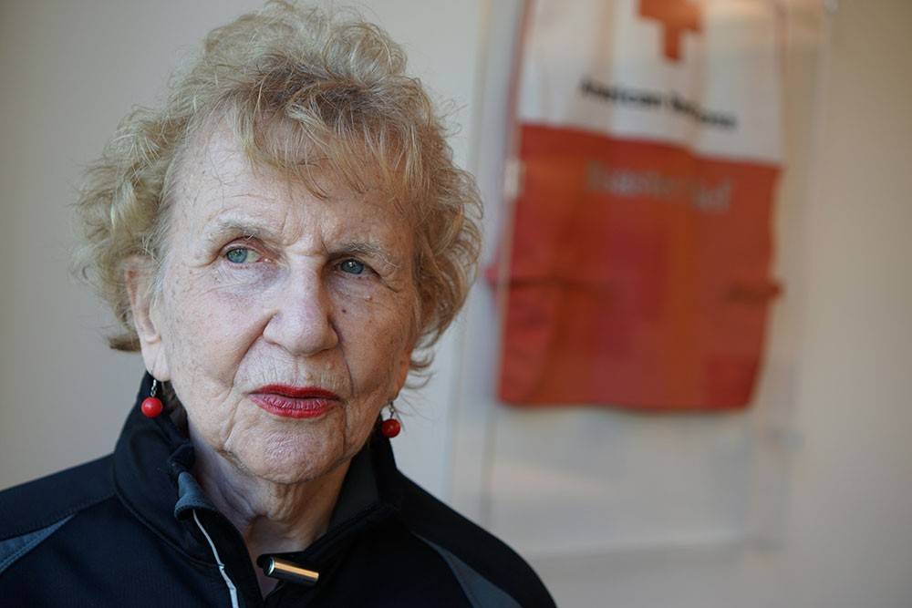 Red Cross volunteer wearing earings with a Red Cross vest hanging on wall in the background.