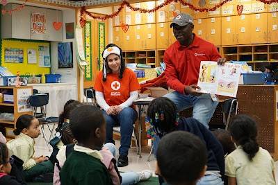 Two Red Cross volunteers wearing hats and Red Cross shirts holding up a book and teaching a class of young students.