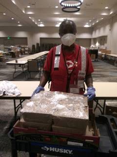 Red Cross volunteer wearing a mask, rubber gloves, a Red Cross vest and shirt inside large event room with boxes, tables and chairs.