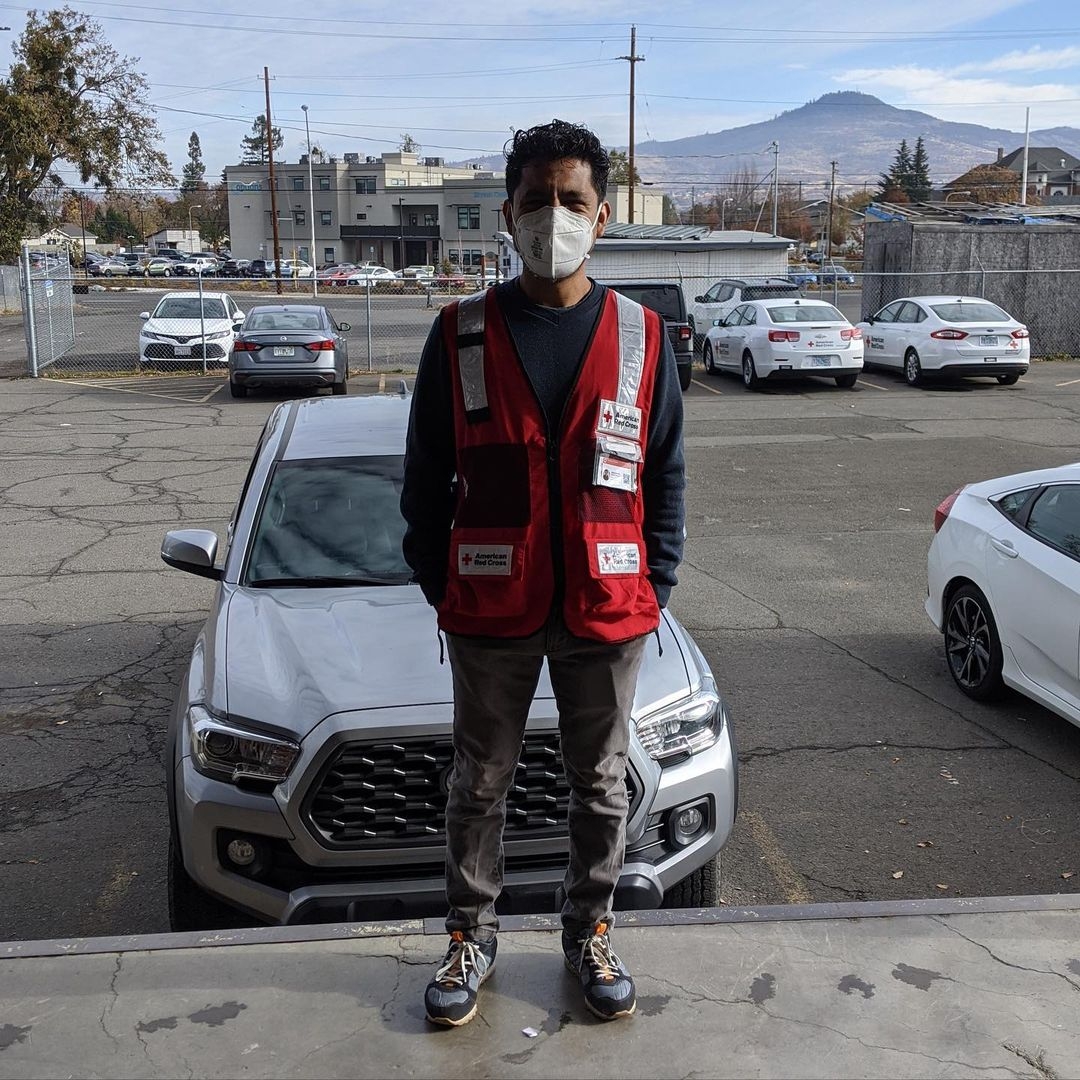 Red Cross volunteer wearing mask and vest standing by parked cars in a parking lot.