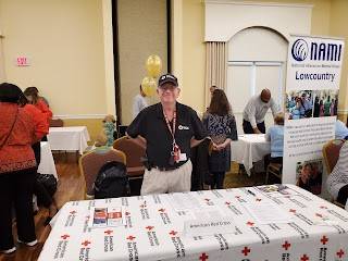 Red Cross volunteer wearing a Red Cross shirt with lanyards around neck standing next to table with Red Cross promotional papers.