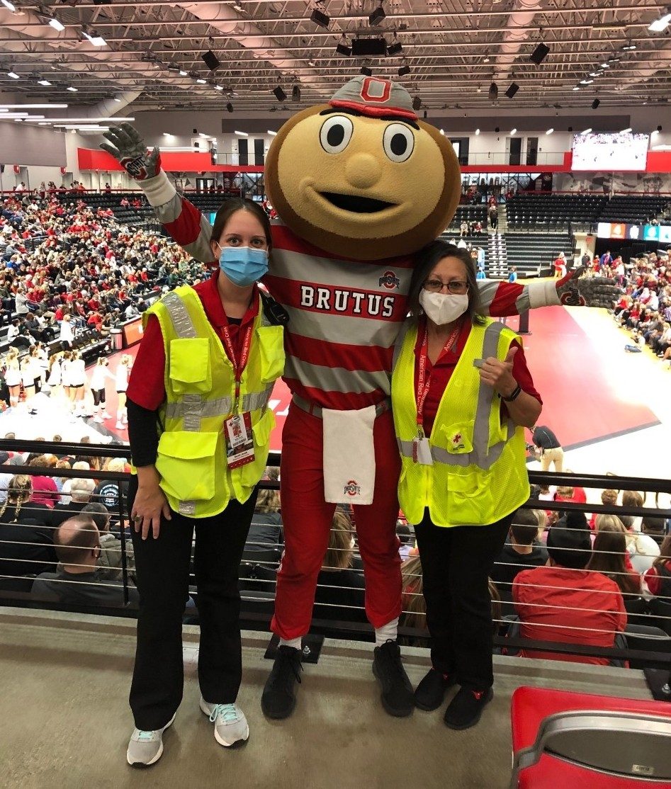 two Red Cross volunteers wearing masks and reflective vests standing next to ohio state university brutus buckeye mascot inside ohio state university basketball court.